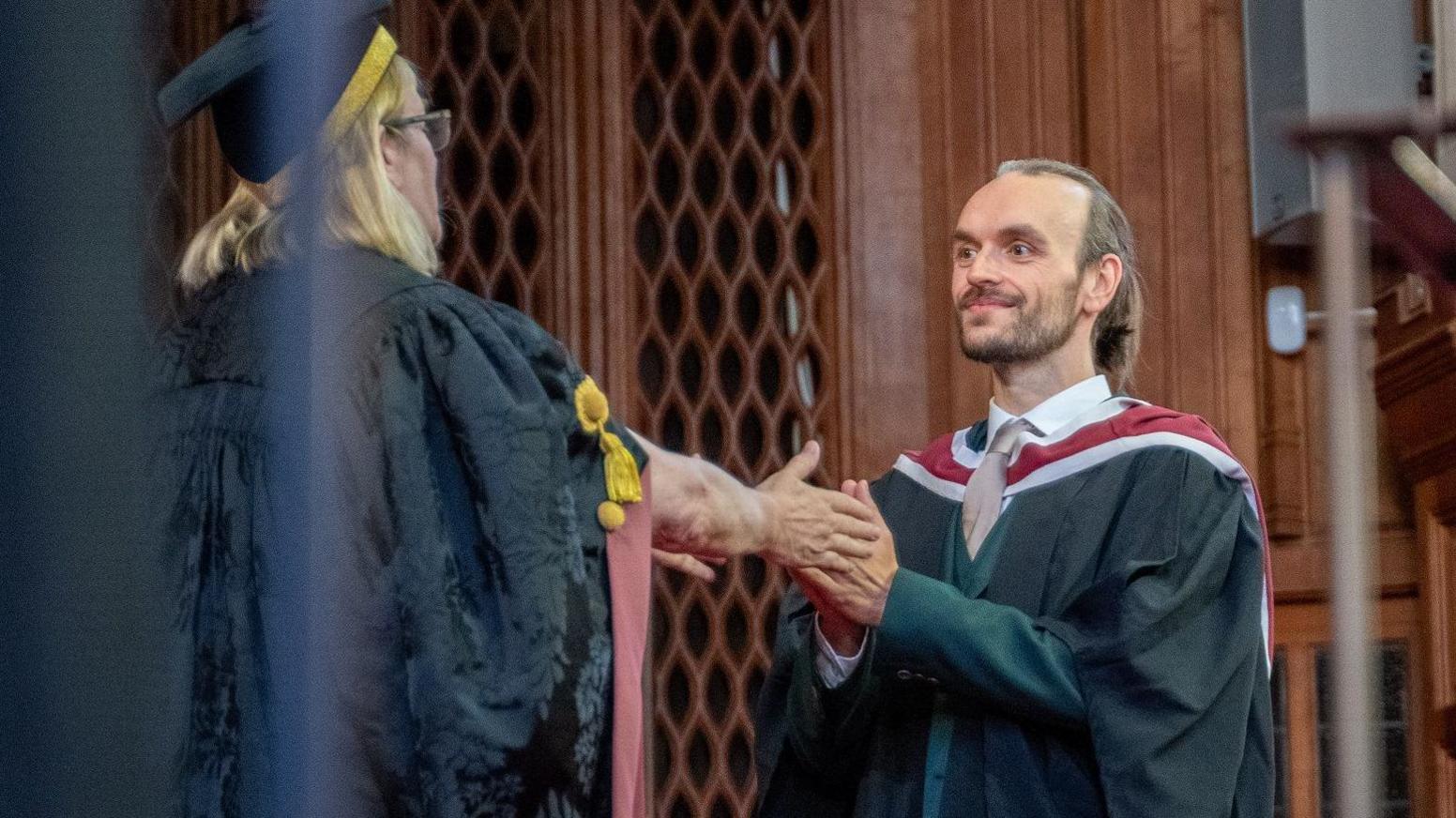 Paul Edwards, who has blonde hair and a beard, wearing gown and standing in hall where he graduated.