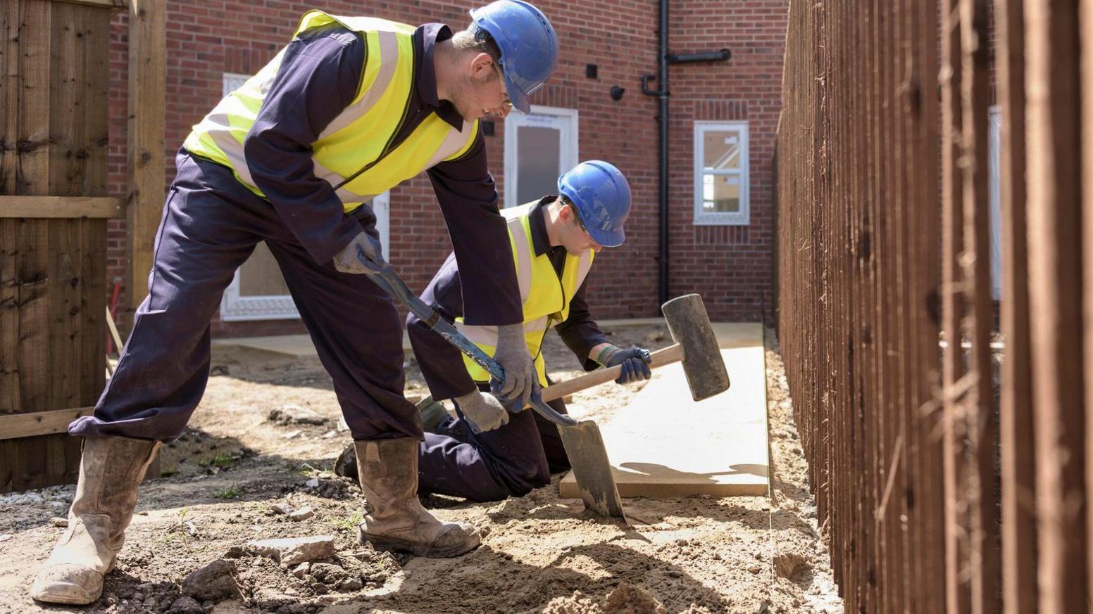 Two men wearing high-vis jackets and helmets digging and hammering on a building site