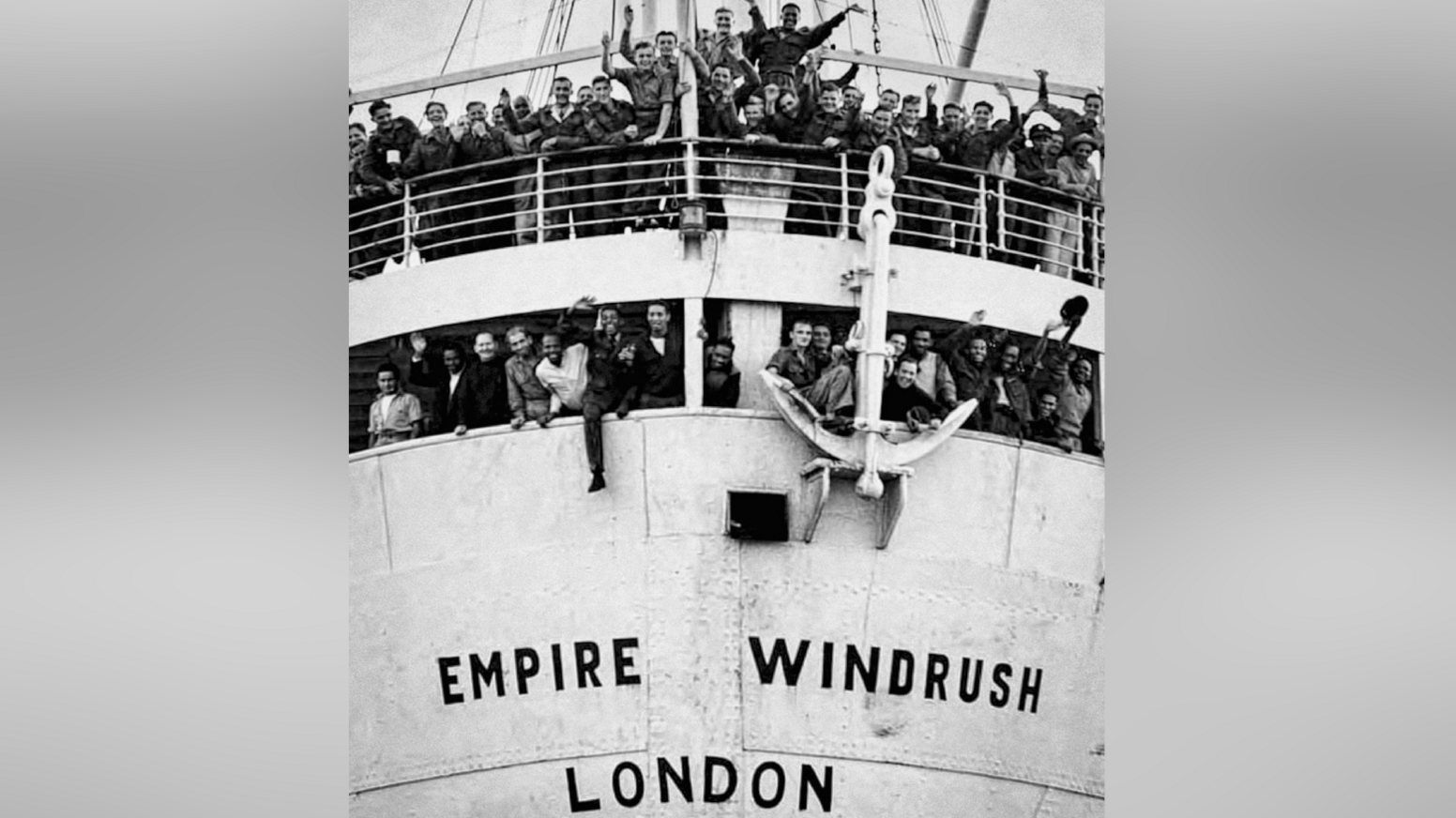 A black and white image of HMT Empire Windrush when it arrived in Tilbury in 1948. Passengers wave and smile from the ship.