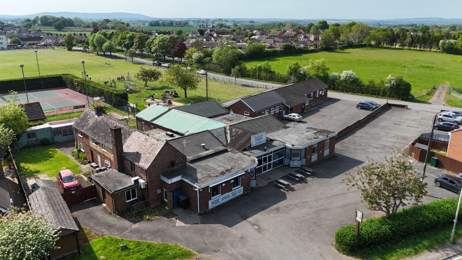 An aerial view of a sprawling one-storey set of buildings with a car park and tennis court alongside and green fields surrounding it
