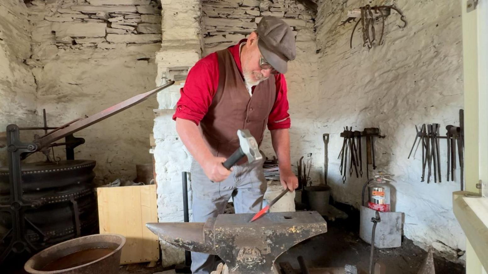 Tony Kornasiewicz is wearing a brown flat cap, has circular glasses on, and is wearing a red shirt with sleeves rolled up, under a brown waistcoat. He's in a small white stone building, surrounded by metal work, and is holding a mallet in one hand and a hot poker in the other, over an anvil.
