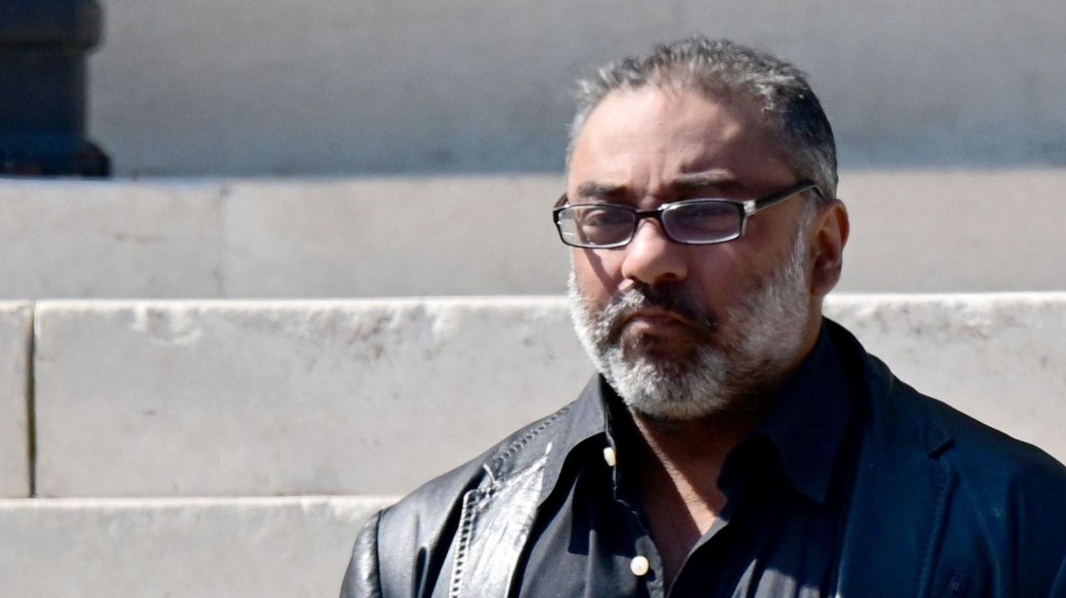 Nadeem standing on the steps of Cardiff Crown court. He has a white beard and grey short hard and rimmed glasses.