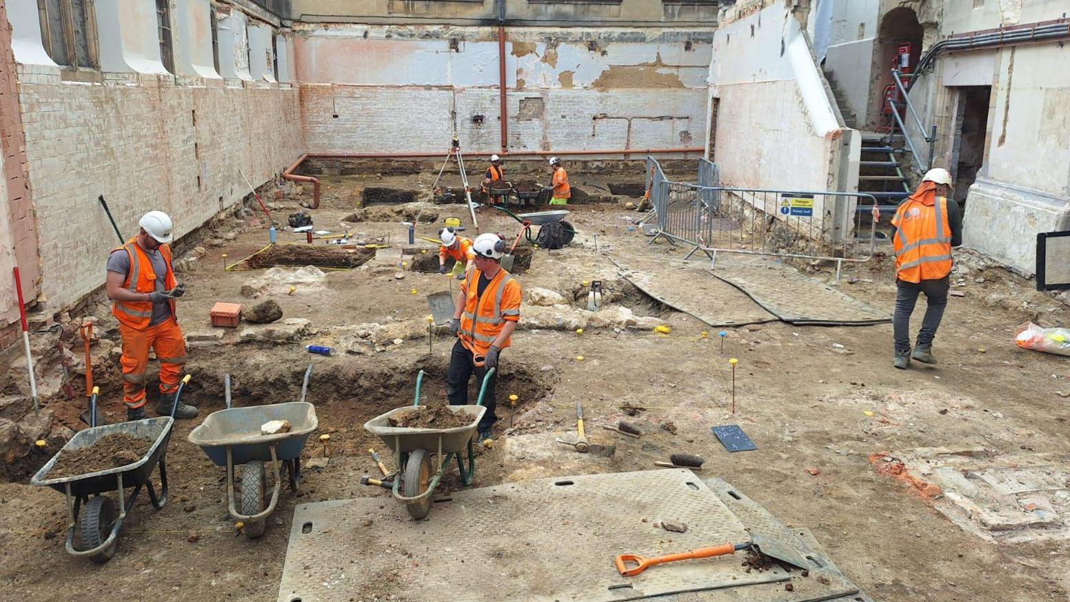 Excavations in the courtyard. There are holes in the ground, people in hard hats and several wheelbarrows.