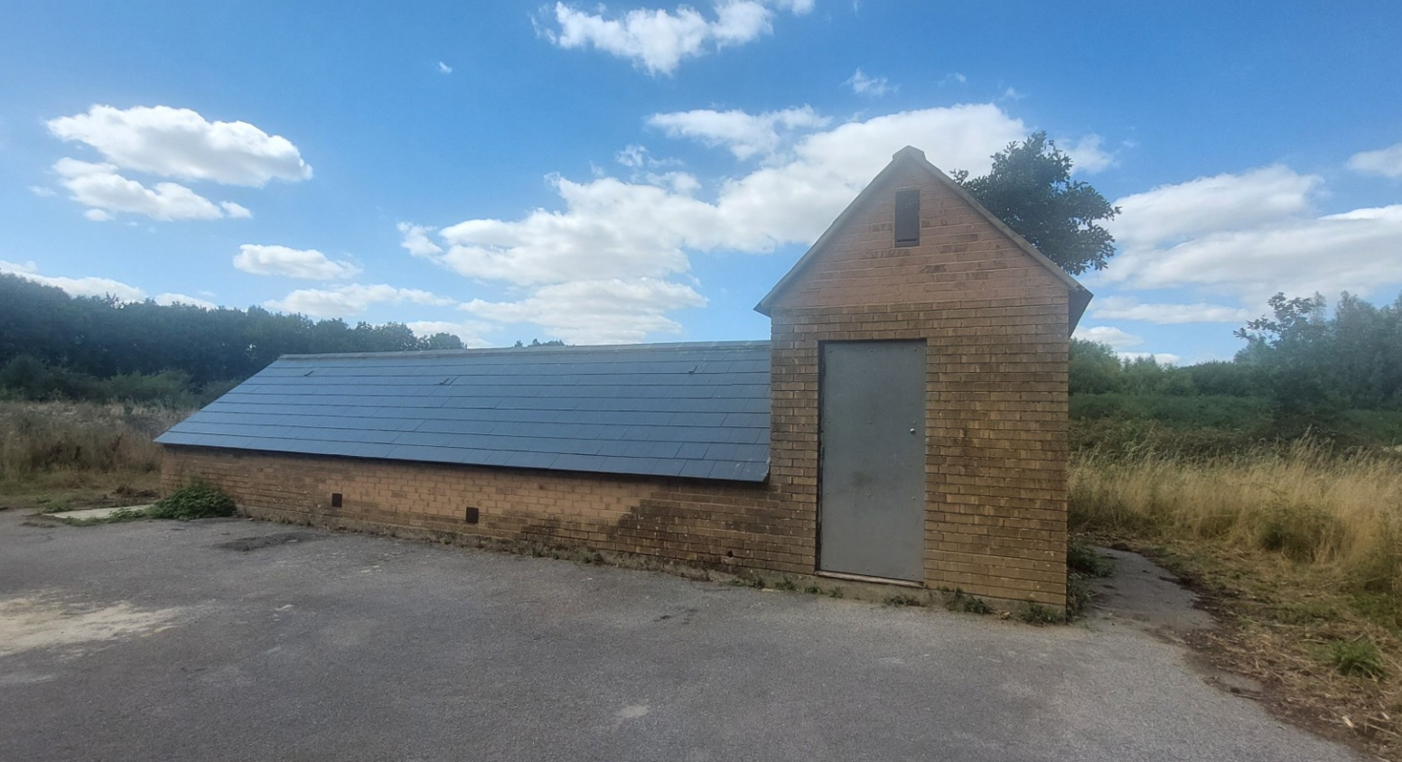 A long brick building that rises only half a storey from the ground with a large pitched tiled roof. The building has a taller section at one end with a grey door, presumably leading to some stairs below ground.