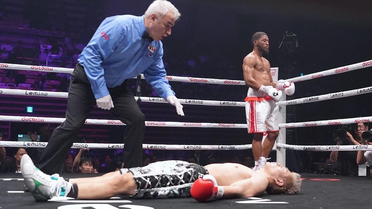 Jin Sasaki lies flat unconscious on his back as the referee moves towards him