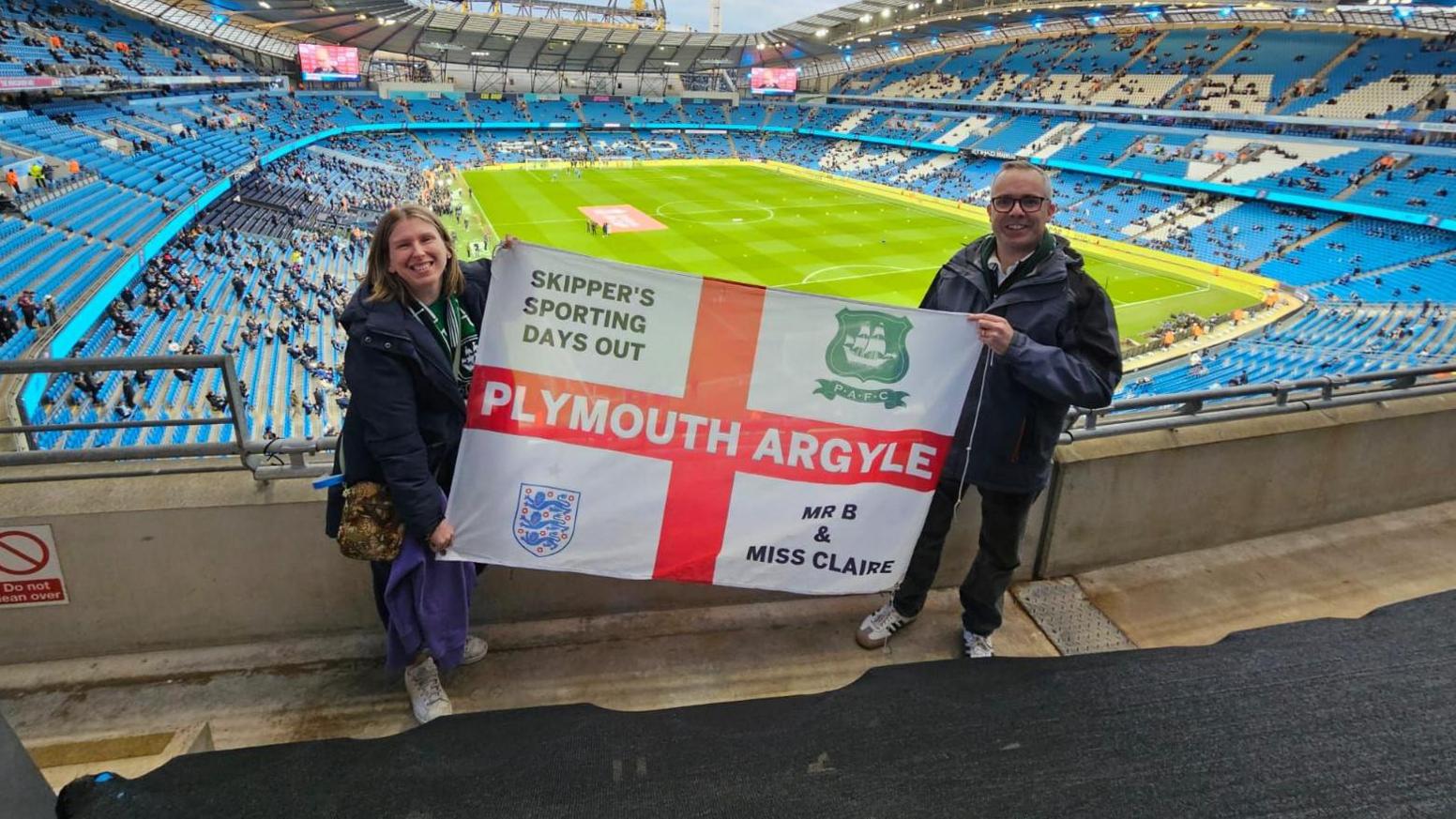 Claire and Tristan Bannister hold an England flag which has Plymouth Argyle written on it along with having Argyle's club crest and the England three lions badge on it. The flag also has messages saying "Skipper's Sporting Days Out" and "Mr B and Miss Claire" written on it. They are standing in the stands of the Etihad Stadium before Argyle's FA Cup match away at Manchester City.