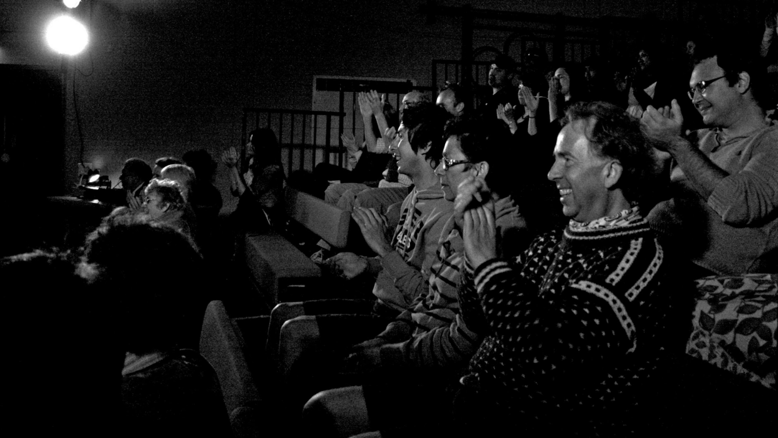 A side profile of an audience sat in a theatre setting. The audience members are sat facing the stage which is out of frame to the left of the image. They are all smiling and clapping. The image is in black and white.