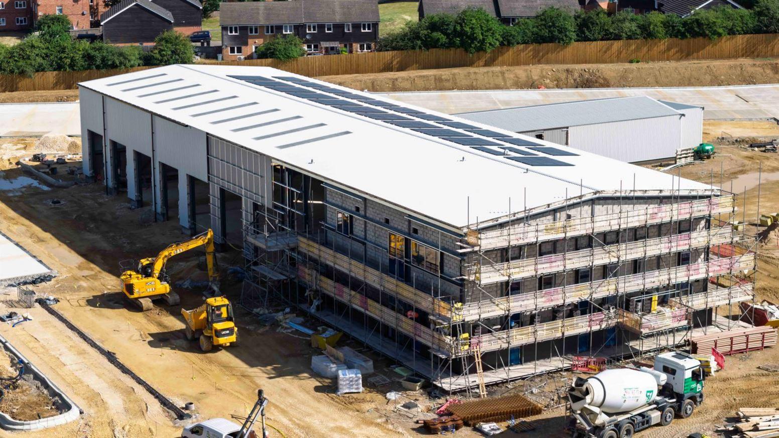 A long rectangular building with three floors is covered in scaffolding and surrounded by a construction site. At the back of it you can see a neighbourhood with several houses.