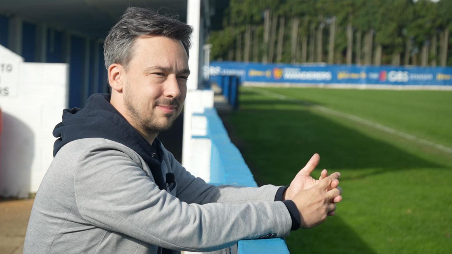Jacques leans over the advertising hoardings, overlooking the pitch at Ipswich Wanderers' Humber Doucy Lane. He has his fingers interlinked as he looks off camera.