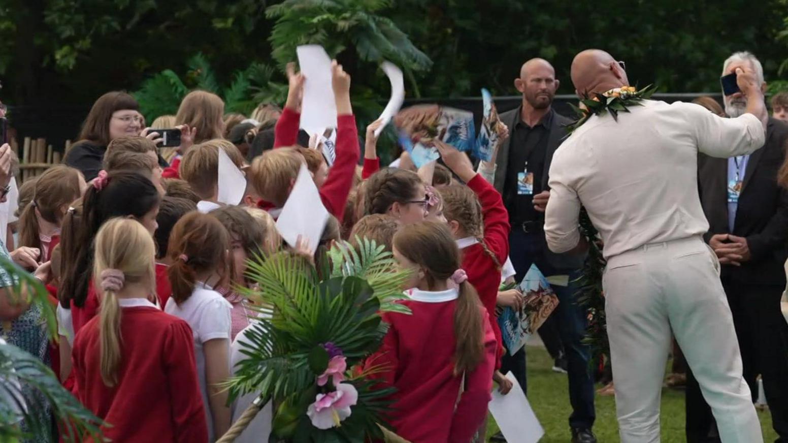 A radical  of schoolchildren wearing reddish  uniforms lasting  successful  beforehand   of the Rock – a antheral   wearing a pick  jumper and achromatic  trousers and sunglasses.