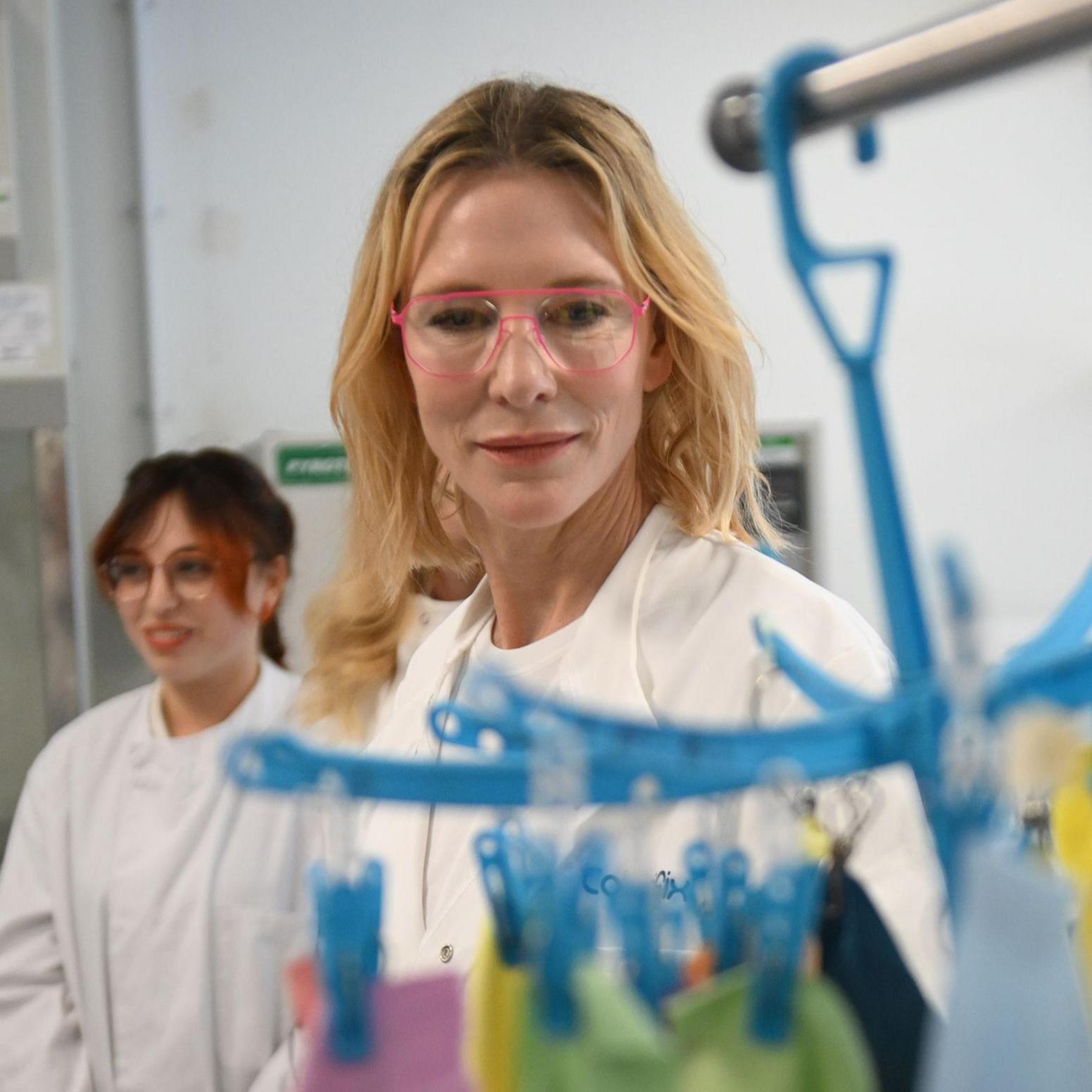 Cate Blanchett a woman who is standing inside a laboratory wearing a white lab coach and glasses.