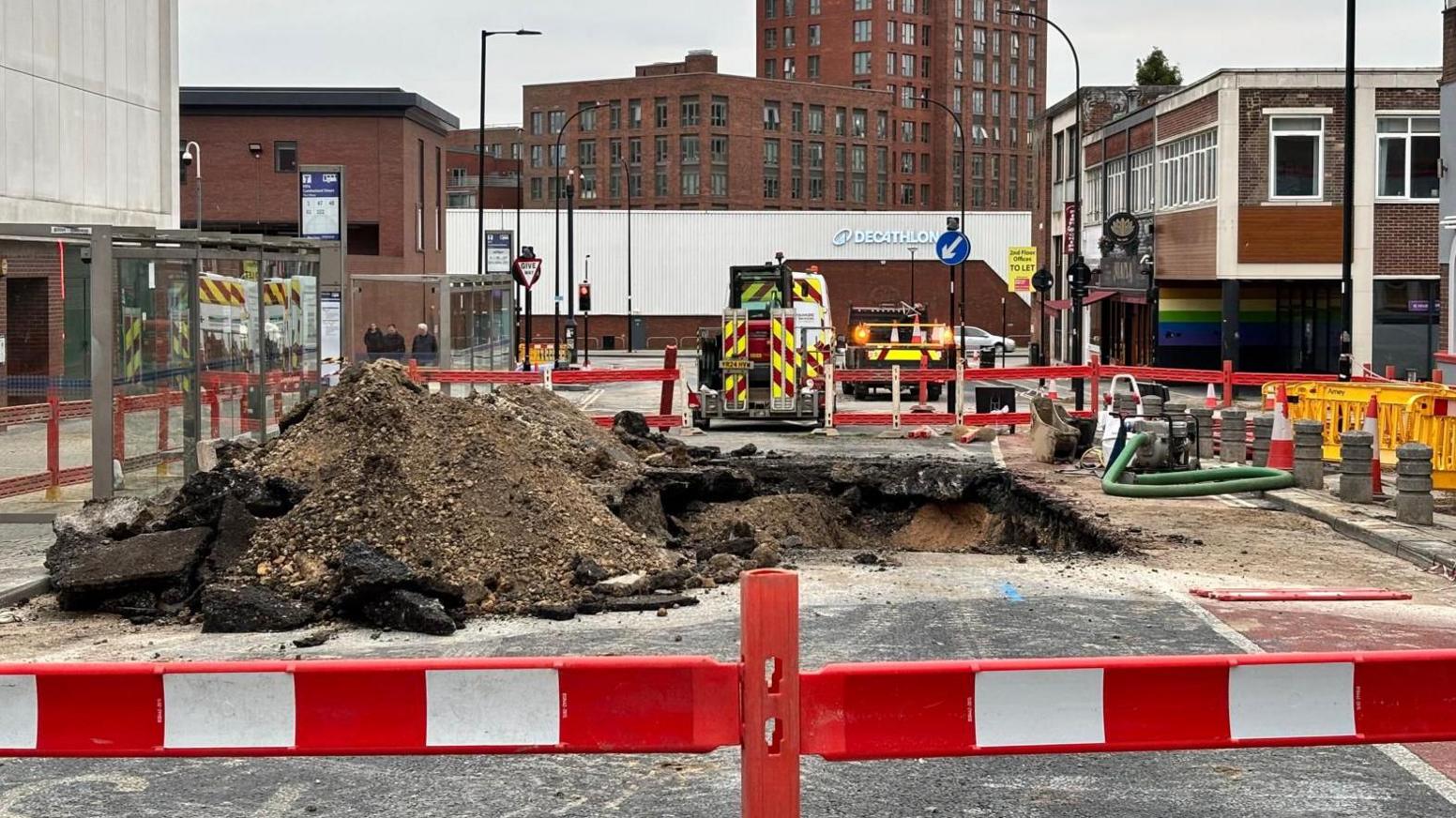 A construction site on street. There is a pile debris in the middle of the street which is cordoned off by red and white barriers. Several construction vehicles and buildings are in the background.