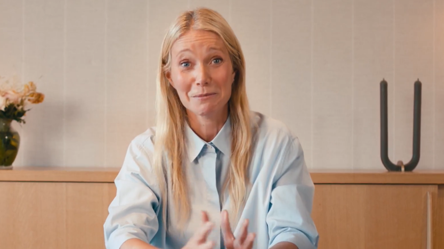 Gwyneth Paltrow appearing in a promotional video for Astronomer. Behind her is a wooden shelf with a plant and a candle on top of it. She is wearing a light blue shirt.