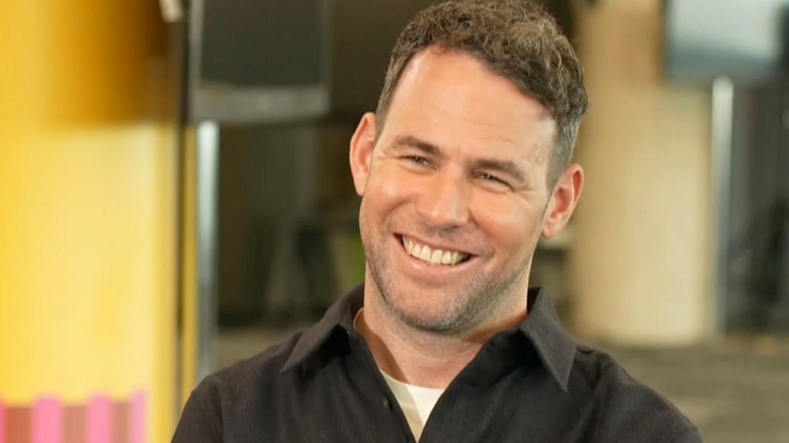 Mark Cavendish is smiling, he has short dark hair, and is wearing a black shirt, with a white t-shirt underneath.
