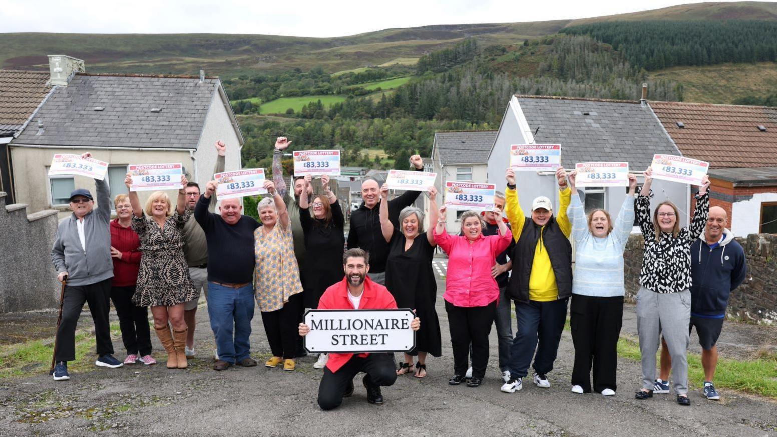 Matt Johnson holding a sign that says 'millionaire street'. He is kneeling on the floor with winners in Nantymoel stood behind him holding their cheques.