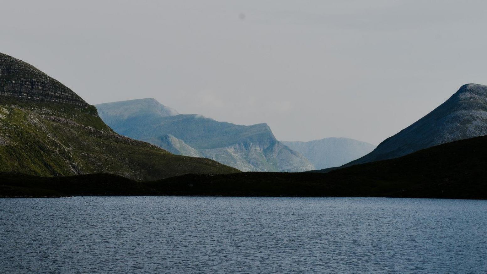 A landscape featuring a calm body of water in the foreground and a range of mountains in the background under a muted sky. 
