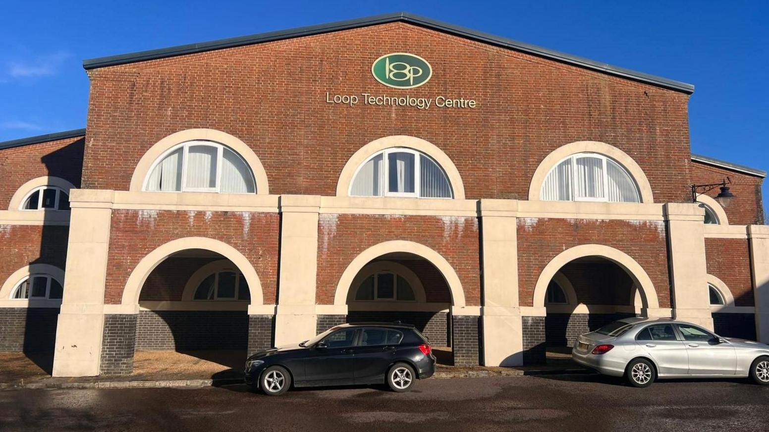 A large red brick building, with a gable-shaped roof, arched windows, and arches at the front. Two cars are parked outside.