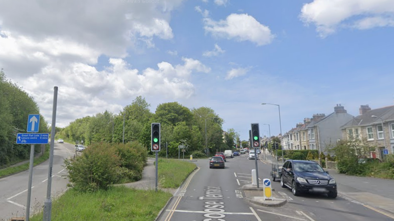 A google street view image of Barncoose Terrace. There is a road going up to the left and a road straight ahead. There is green grass and a pedestrian footpath in the middle of the two. There are cars on the road driving in both directions. There are traffic lights which are green. There are houses on the right.