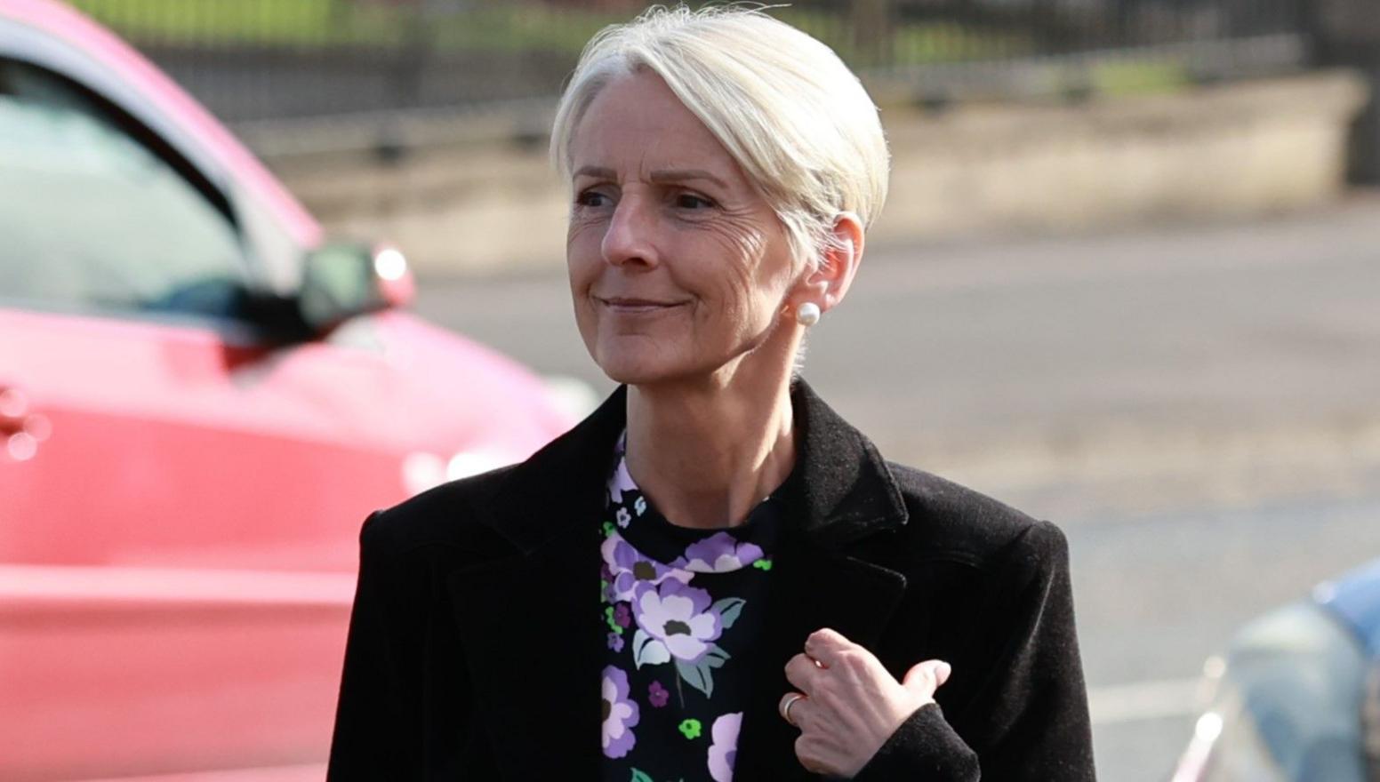 Lady Donaldson with short light hair, wearing a black jacket and purple flowery top.