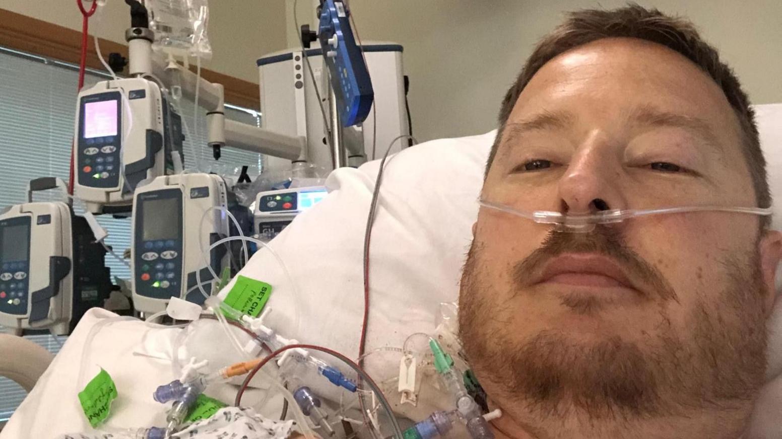 Man in a hospital bed, wearing a hospital gown, with lots of wires connected to monitoring equipment by his bed.