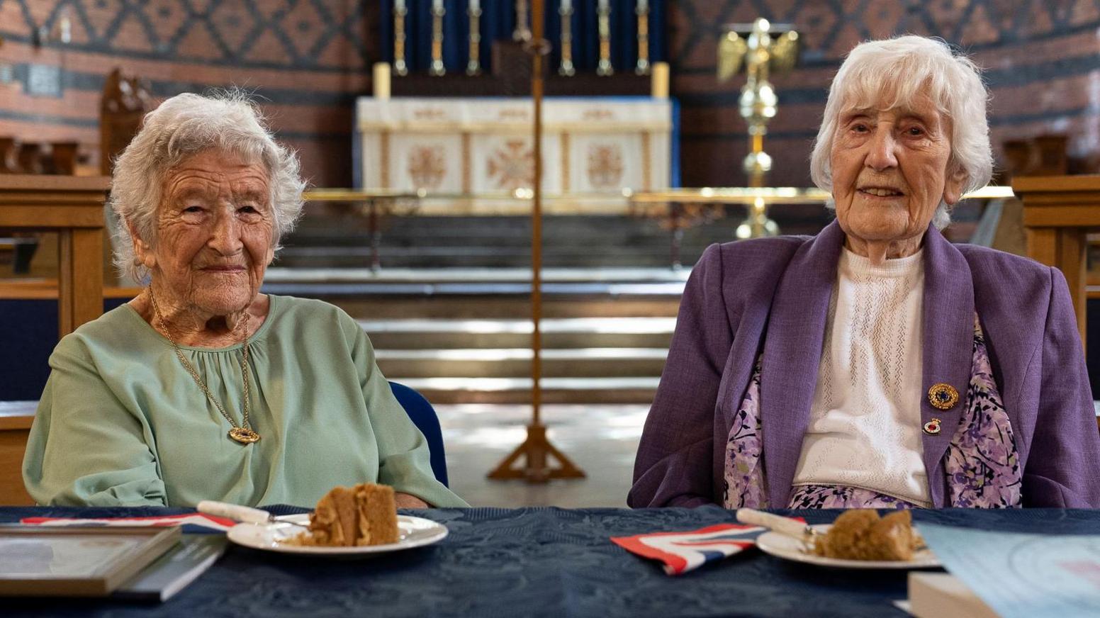 The two codebreaking ladies sitting in the church where they have been presented with an award. Kath is wearing a green top while Gwenfron is wearing a purple jacket, both have short white hair. There is cake laid on the table along with a framed a certificate and a book.