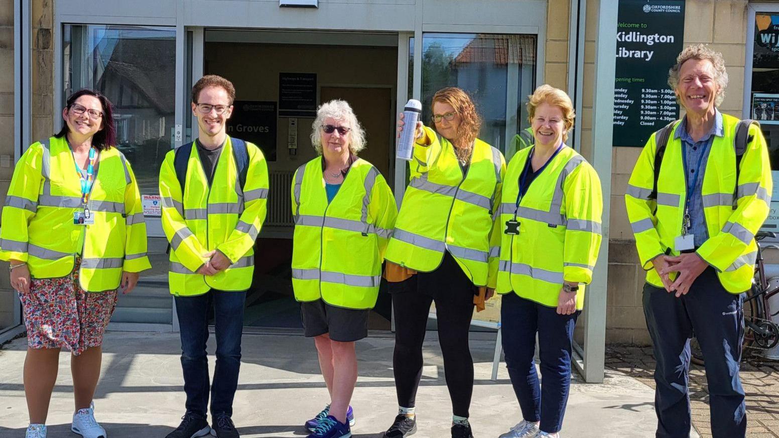 Six FixMyStreet superusers, all dressed in vis-vis jackets, standing outside Kidlington Library, including a woman with mid-length ginger hair, who is showing the person taking the picture a spray can of paint 