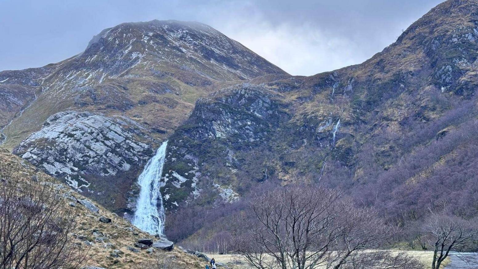 The waterfall rushes down a steep mountainside. Mountain summits rise about the rushing white water. 