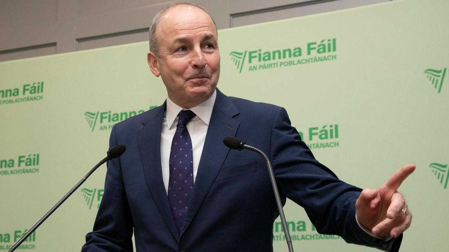 Micheal Martin who has thinning light coloured hair and is wearing a navy suit and tie with a white shirt looks to his left and points to someone while standing on a podium with Fianna Fail signs in the background