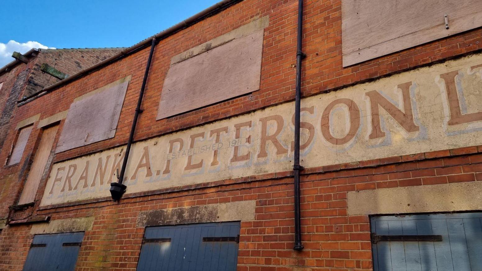 A derelict brick building with the sign Frank A Peterson on the front. Windows on the second tier are boarded up. There are blue wooden doors on the bottom level