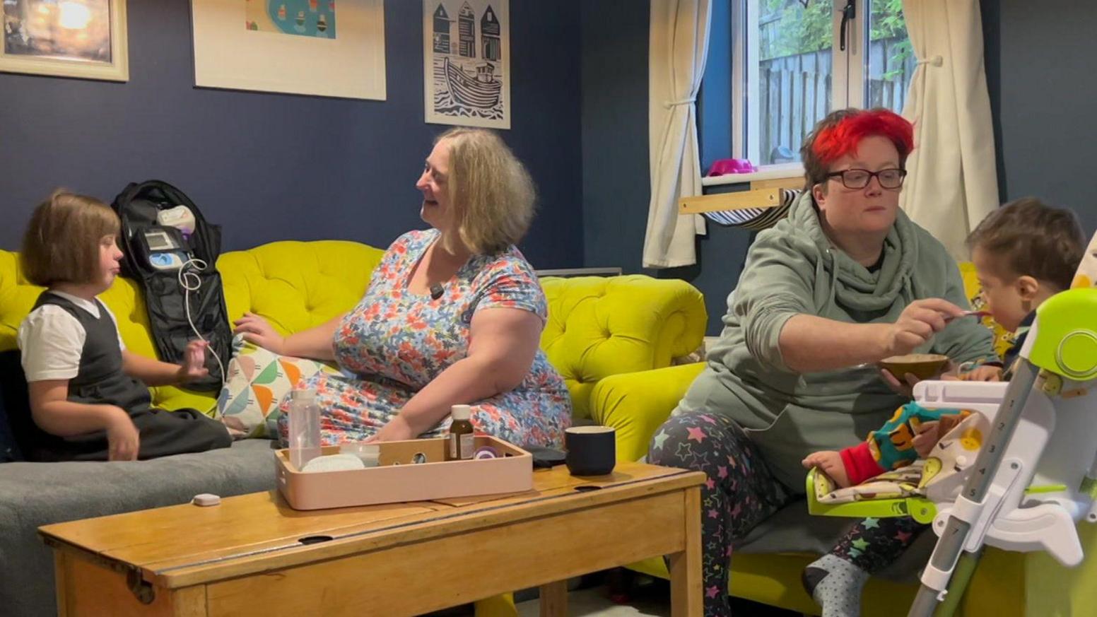 A family of four in their home. There are two adults and two children. The adults are feeding the young children. They are in their living room which has bright yellow sofas and dark blue walls with lots of picture frames on the wall and white curtains next to a window. 