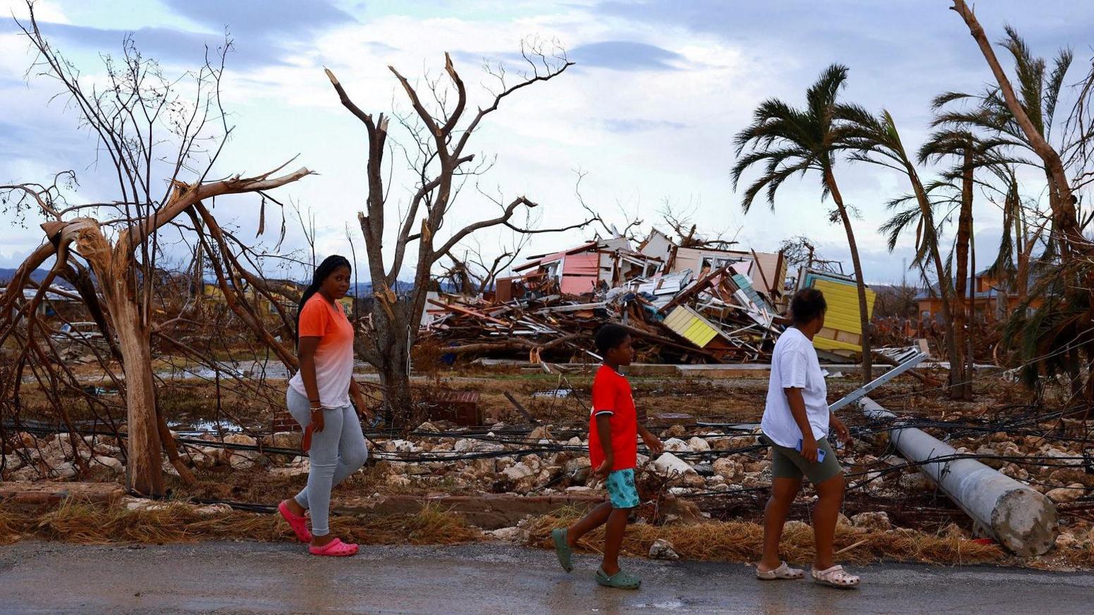 A young woman, boy and an adult walk past widespread debris and damaged trees. A mound of collapsed housing is in the background.