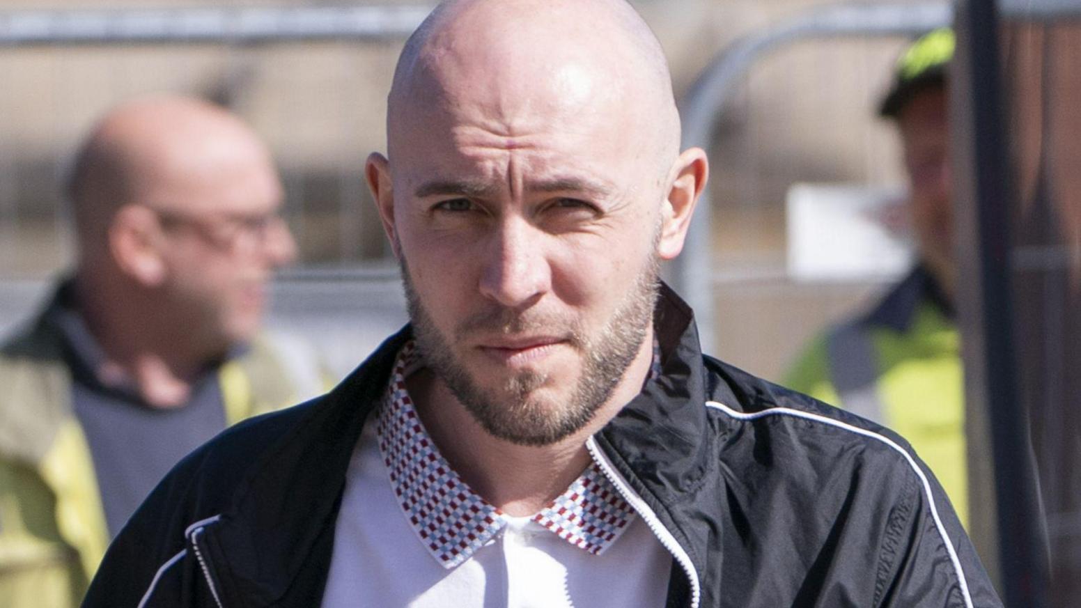 Daniel Flatters, who has a bald head and dark stubble, looks into the camera with a serious expression