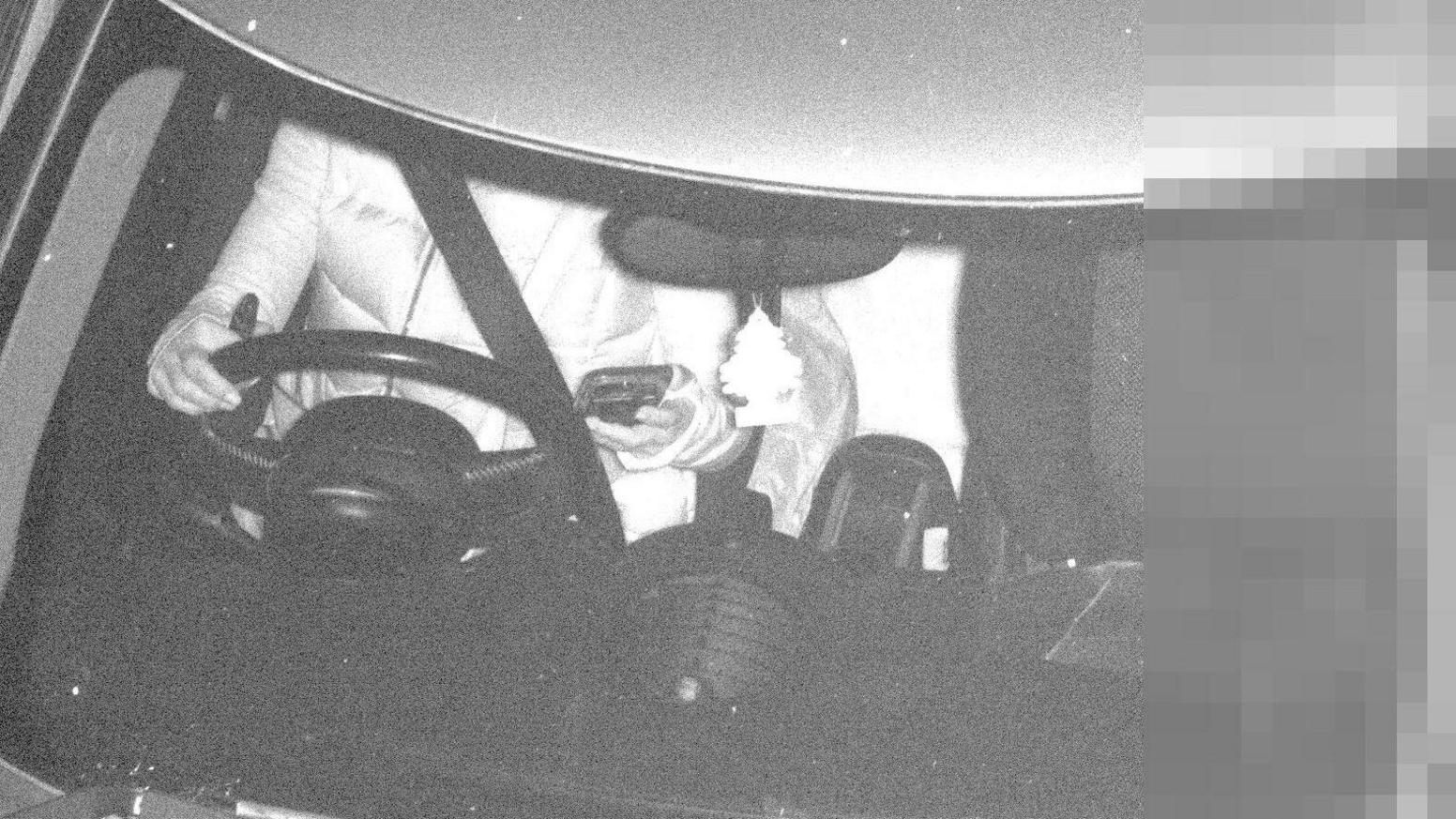 A black and white picture looking into a car windscreen. The driver, who is unidentifiable, has one hand on the wheel and another holding a mobile phone