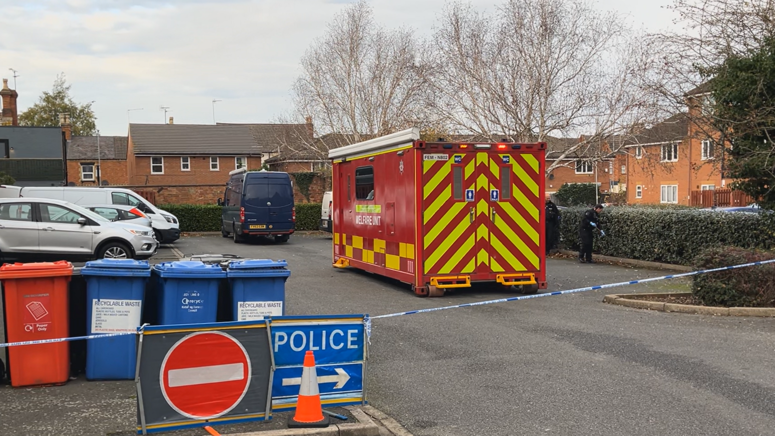 A red welfare unit is parked in a car park outside a block of flats. A police cordon blocks off that area and there is a blue sign that reads police with an arrow pointing to the right. Several bins rest together to the left of the image within the cordon. Some vehicles are parked within the cordon.