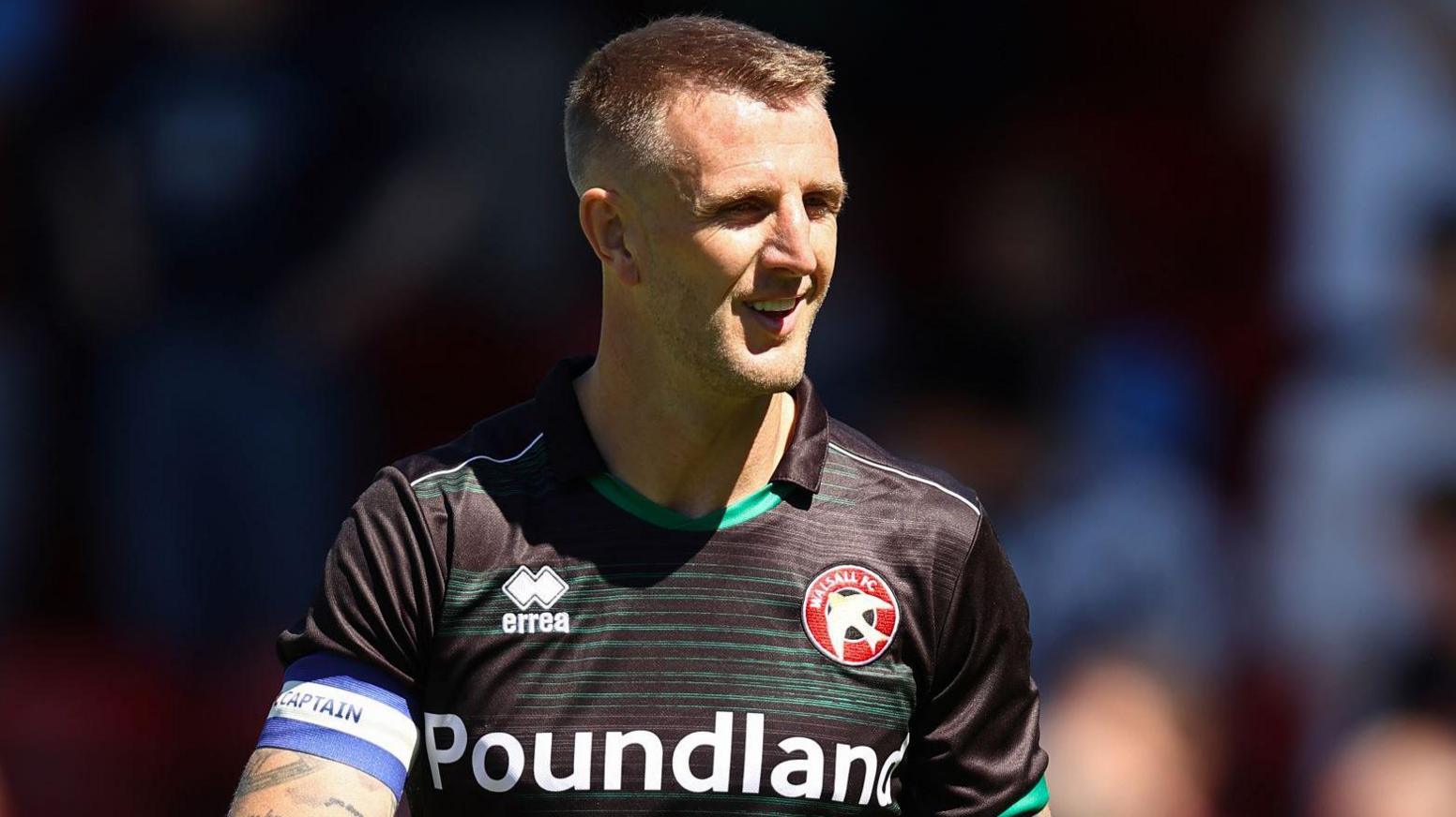 Peter Clarke wearing a black Walsall football top while on the pitch. He is squinting and smiling. He is looking away from the camera.