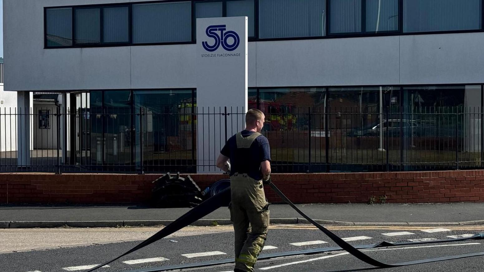 A man wearing a fire kit holding a large hosepipe outside a building.