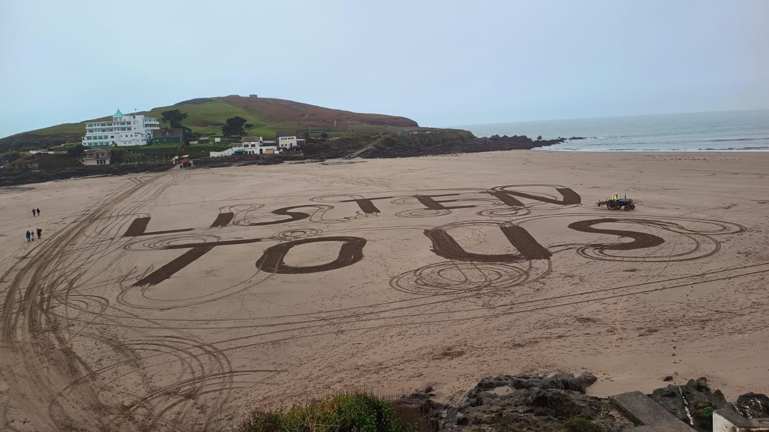 An elevated view of Bigbury Beach which has the words "Listen to us" written in giant capital letters in the sand. The beach is otherwise empty.