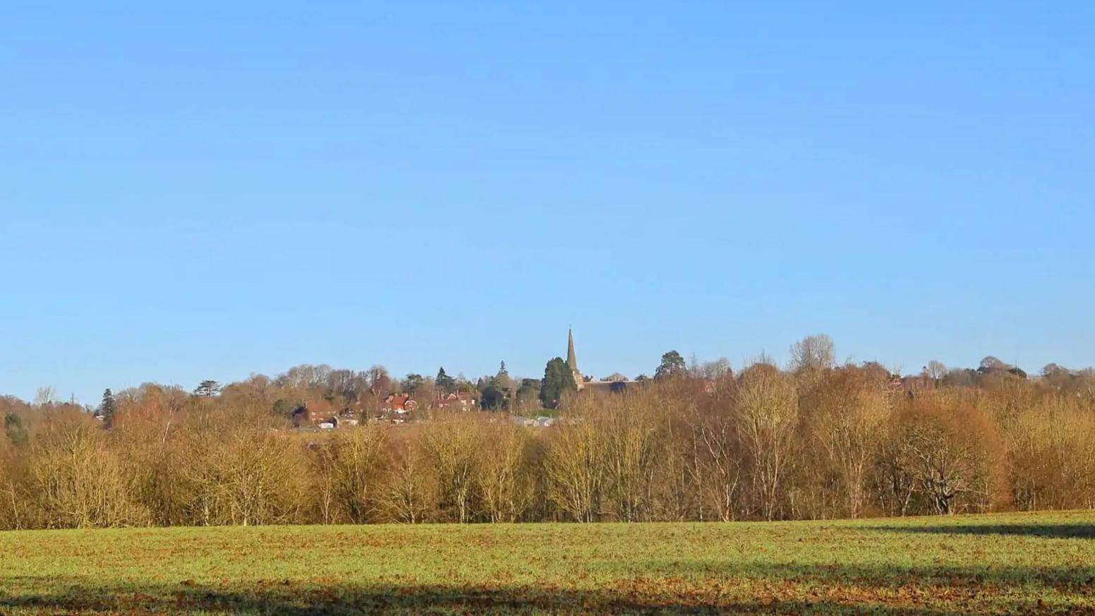 A shot of rural land showing a field, woods and a village in the distance with church spire on a sunny day.