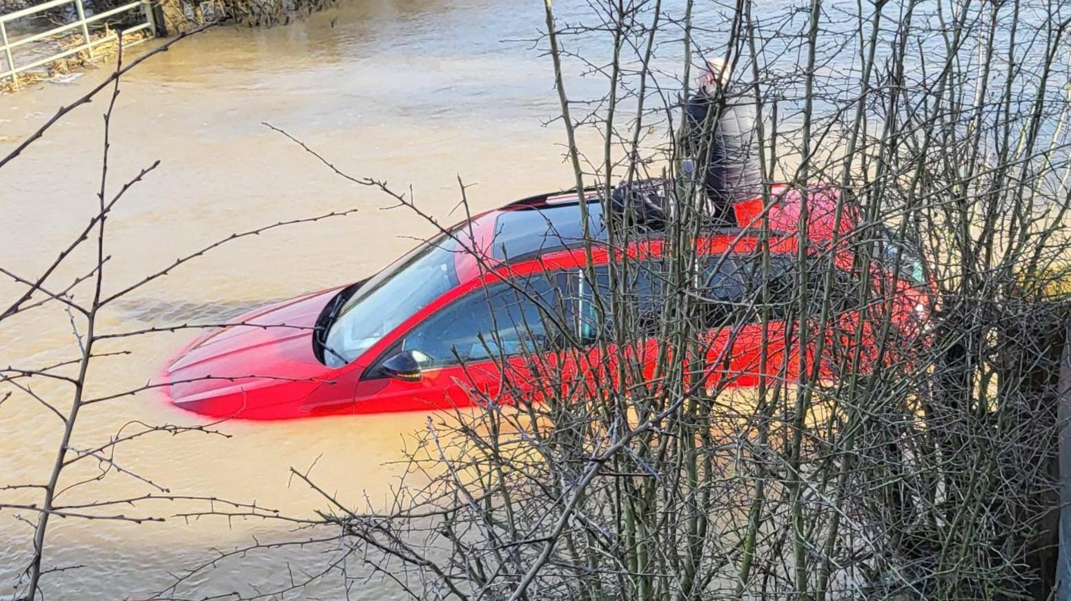 A red car immersed in flood water with a person sat on top of it with a bag beside them, obscured by trees