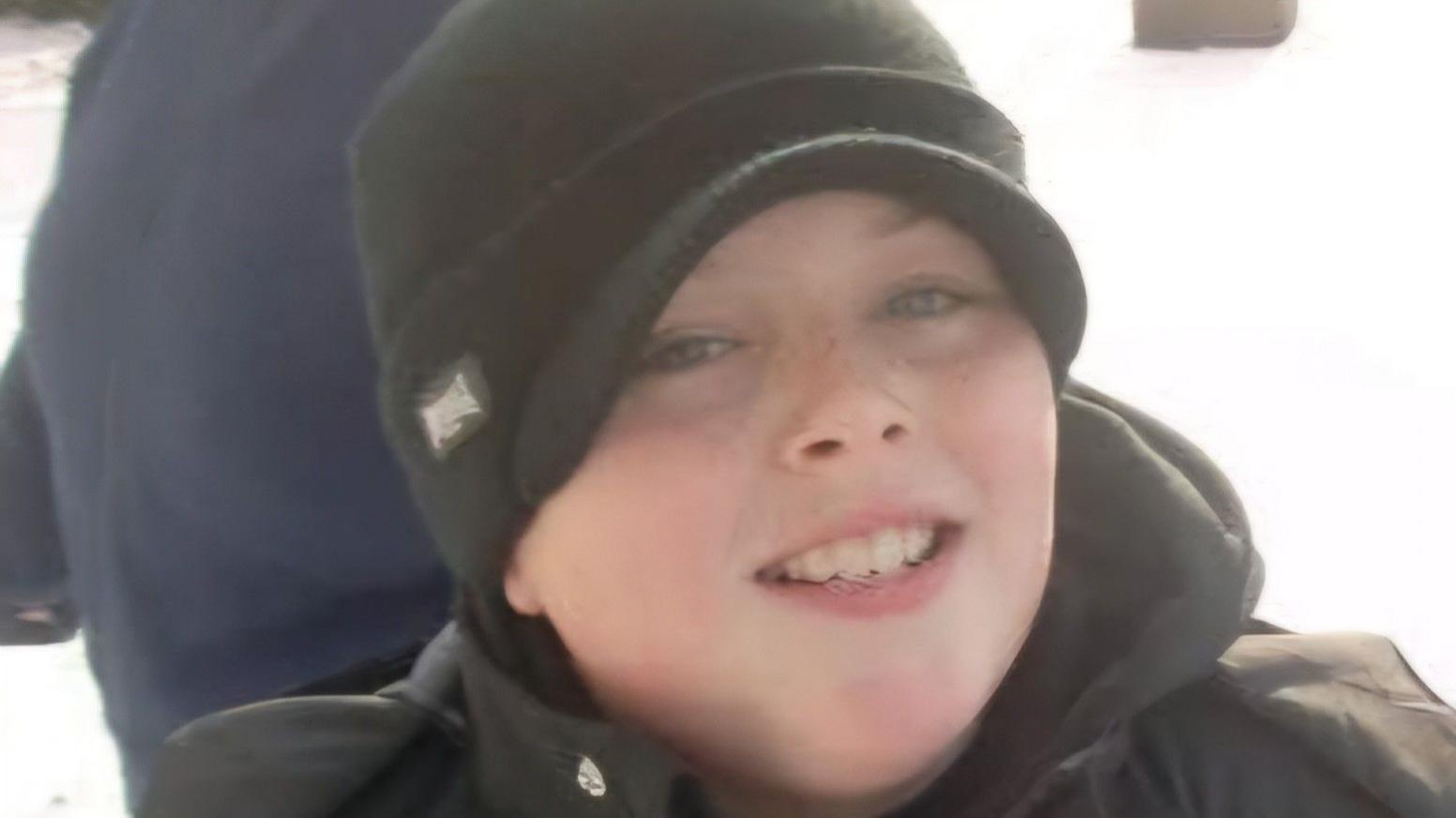 Connor, aged about 12, wearing a black coat and black beanie hat smiles at the camera. He appears to be outside, in the snow.
