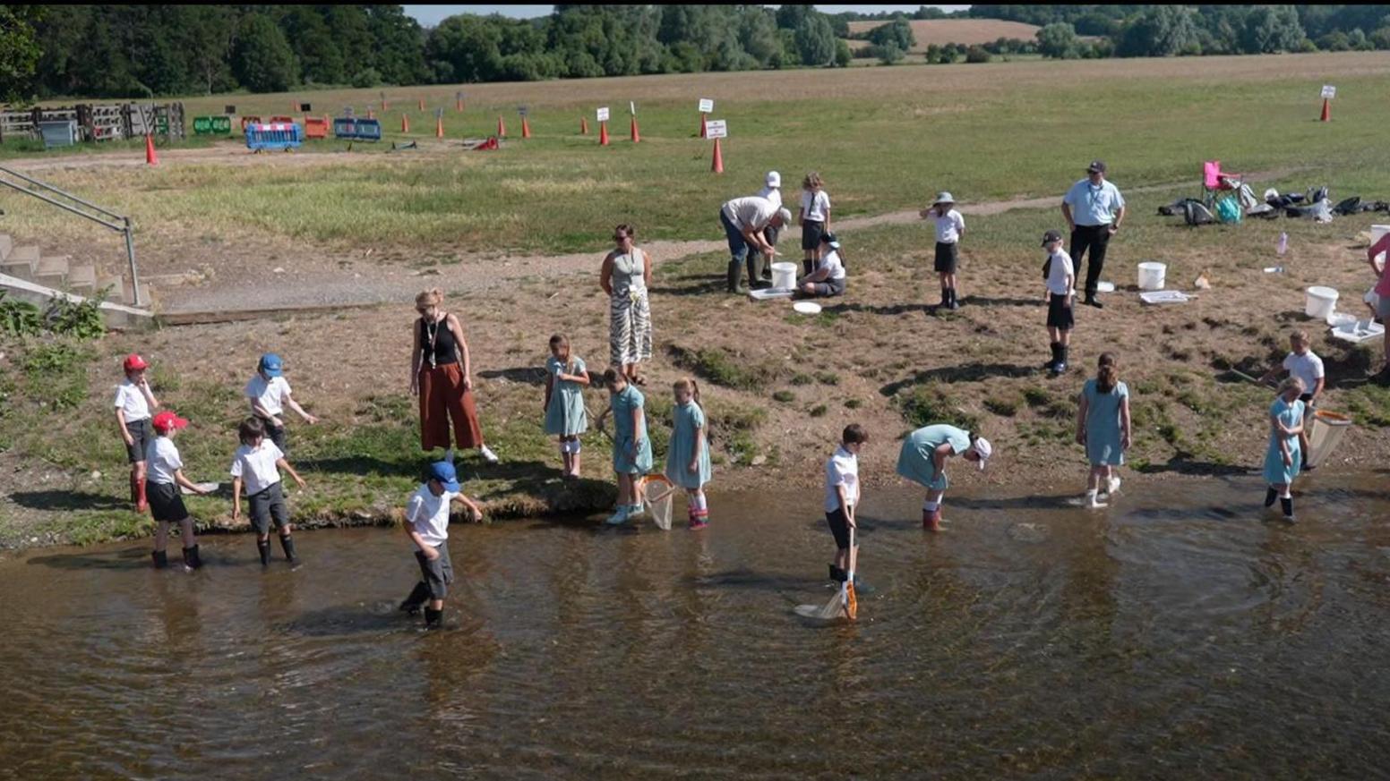 A group of children stand in a river wearing their school uniforms while carrying out water sampling using nets and trays