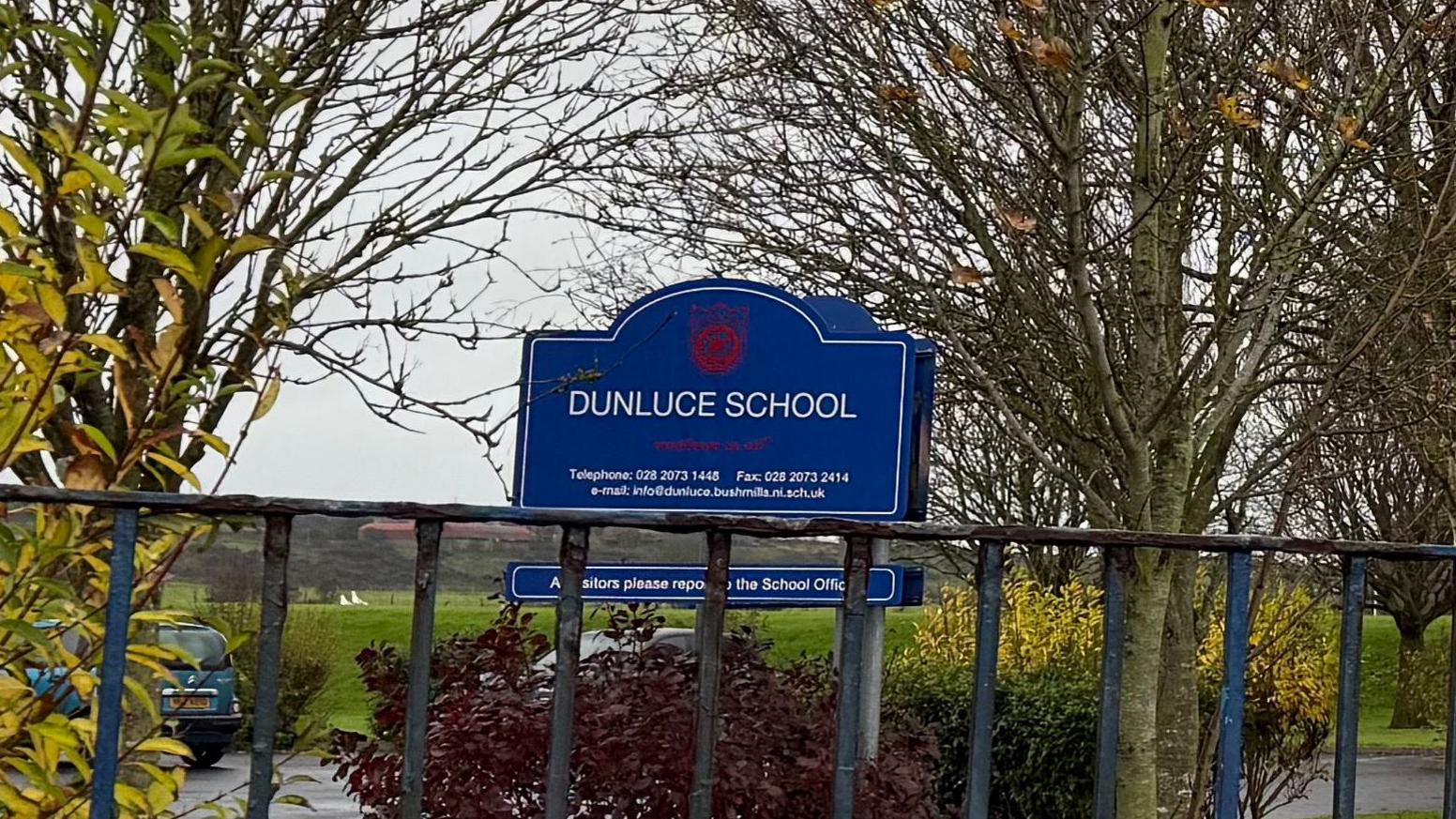 A blue sign which says 'Dunluce School' with a red school logo above and contact information beneath it. There is metal fencing in front of the sign and trees around it.