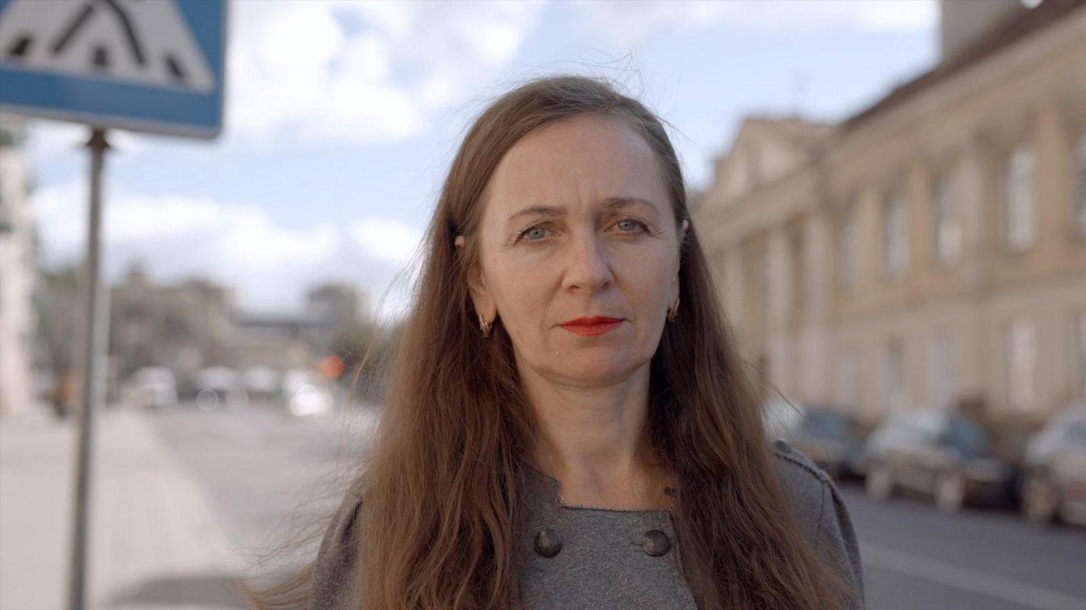 A woman with long brown hair stands in a street and stares at a camera
