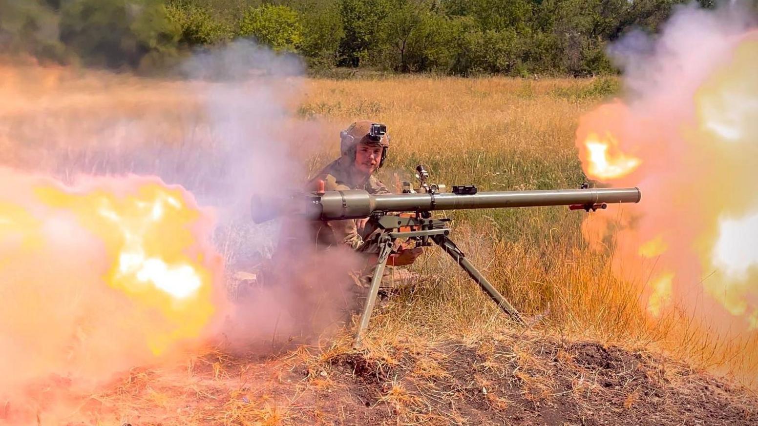 James Scott Rhys Anderson kneeling down firing a rocket from a launcher with fire on either side.