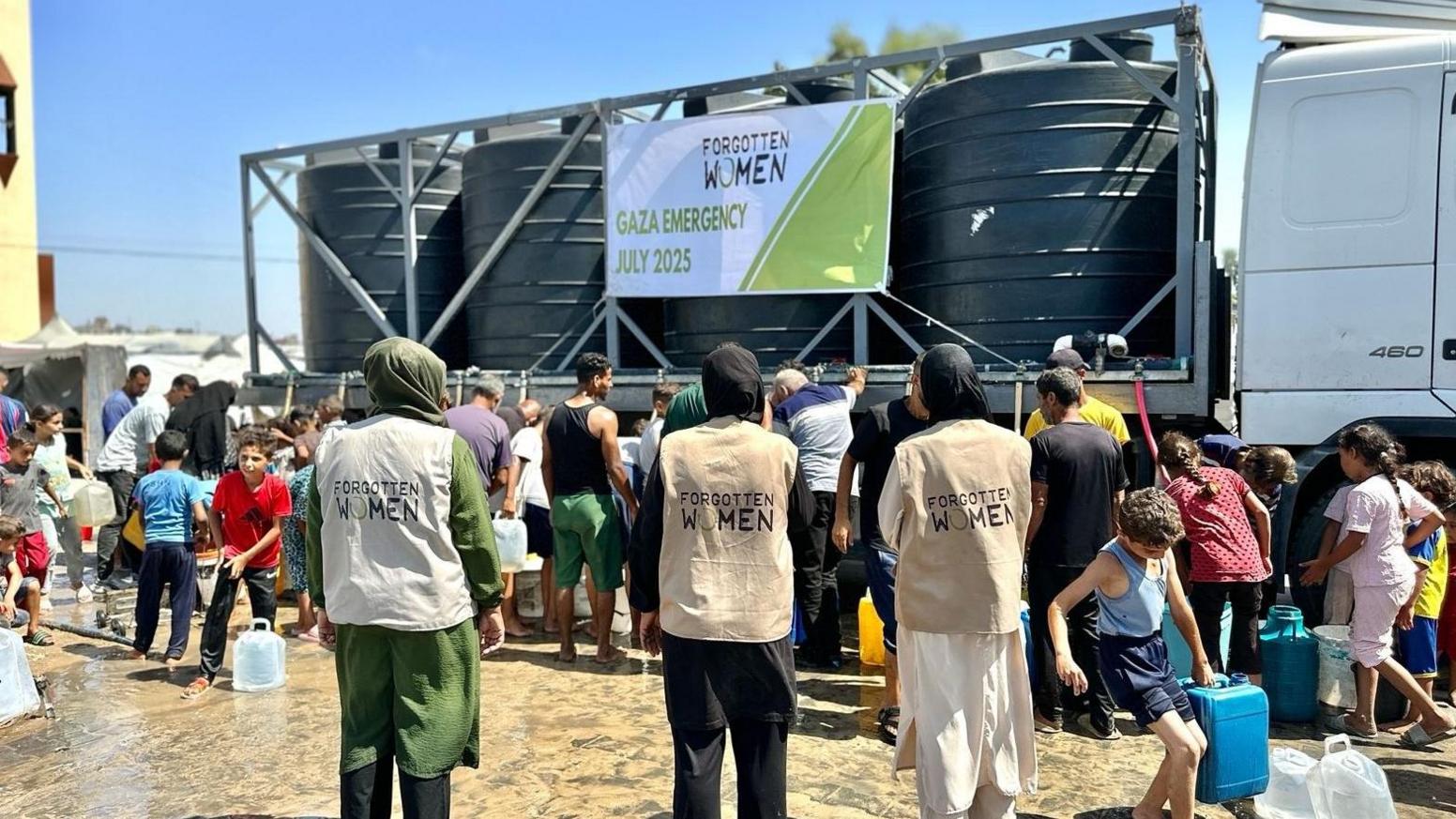 Crowds of people stand in front of a van with large black barrels on the back. Several women in hijabs and jackets with "Forgotten Women" on the back stand at the back.