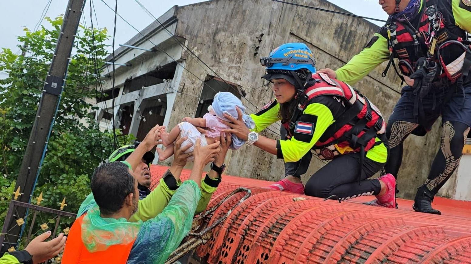 Rescuers on top of a roof, passing down a baby to other rescuers on a boat in floodwater