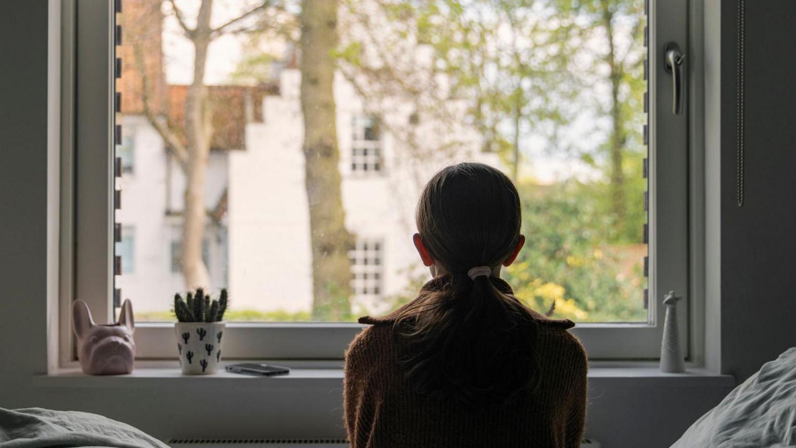 A young girl looks out of her bedroom window. The view through the window is blurry but shows a residential area.