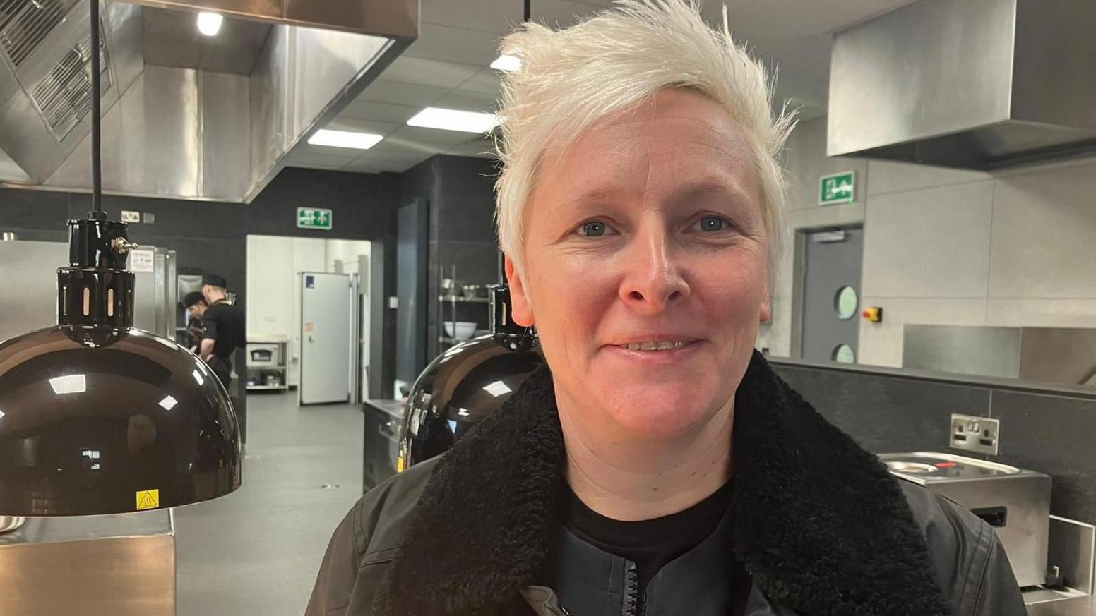 Lisa Goodwin-Allen in the new facility at Blackburn College. She stands in the kitchen by a hot plate and various other appliances. Lisa has short bleached blonde hair and is wearing a black jacket.