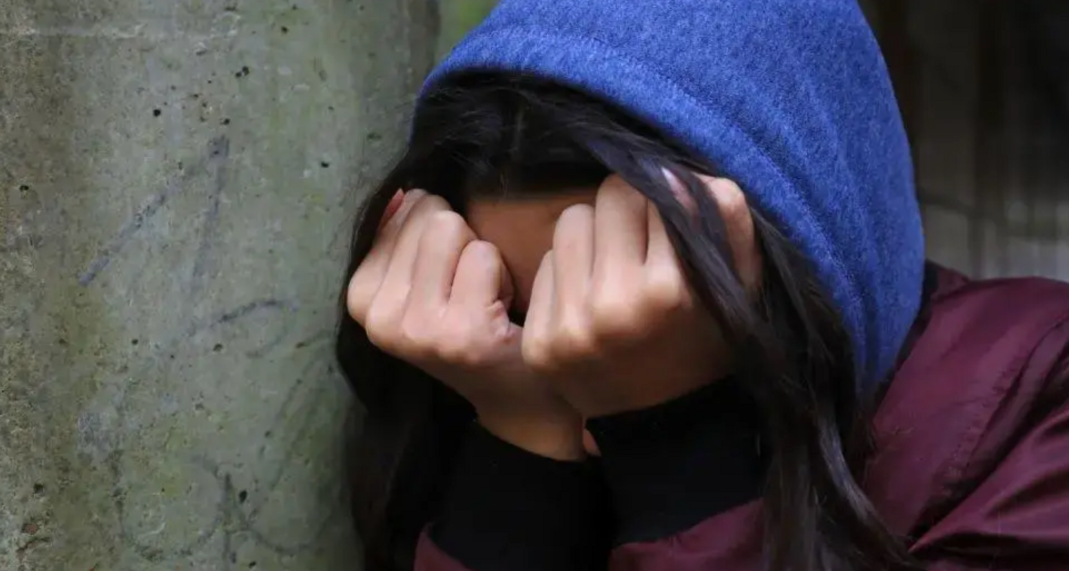 Person with long black hair wears blue hoodie and burgundy coat. Standing against a concrete wall the person cover's their face with their fists in a distressed manner.