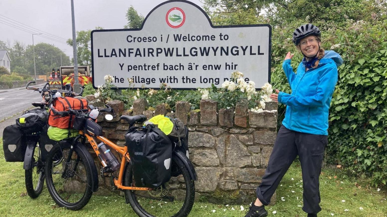 Becky Sherwood wearing a blue coat and helmet. She is standing next to a sign that says 'Welcome to Llanfairpwllgwyngyll'.